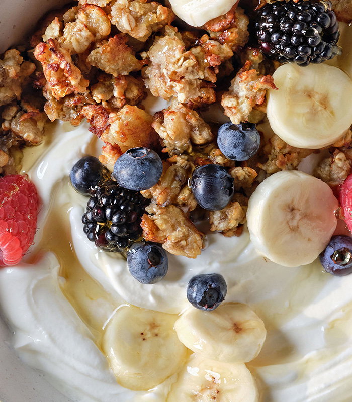 A bowl of creamy yogurt topped with granola, bananas, blueberries, blackberries, raspberries, and a drizzle of honey.