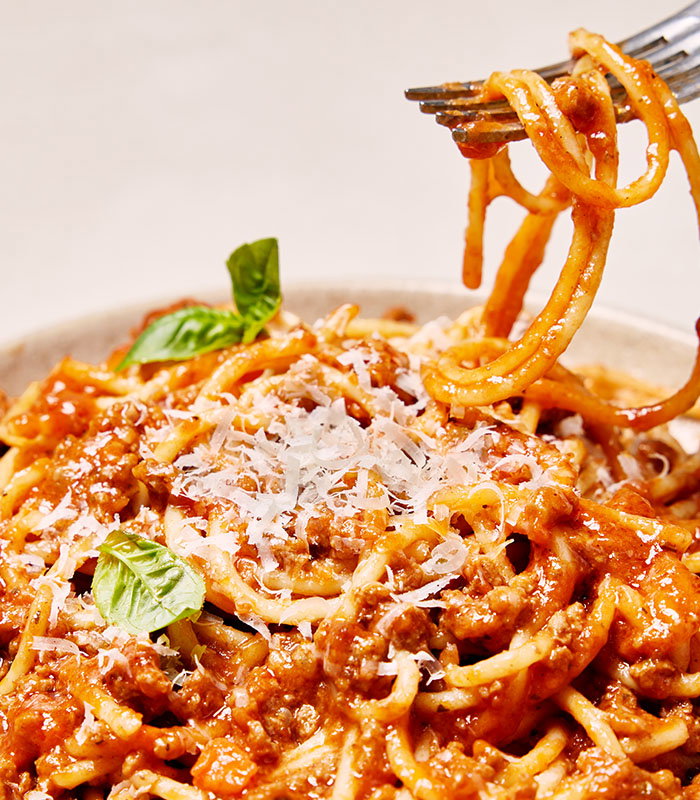 A fork lifts spaghetti topped with meat sauce and grated cheese, garnished with fresh basil, against a beige background.