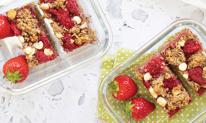 Two glass containers hold raspberry oat bars topped with white chocolate chips, surrounded by fresh strawberries on a green polka dot napkin.