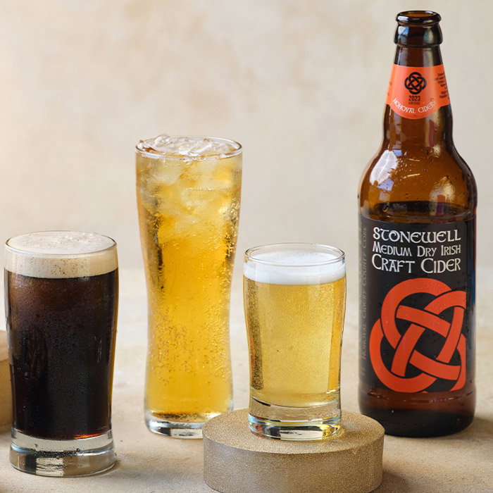 Three glasses of different beverages, including dark beer, a tall iced drink, a light beer, and a bottle of Stonewell craft cider.