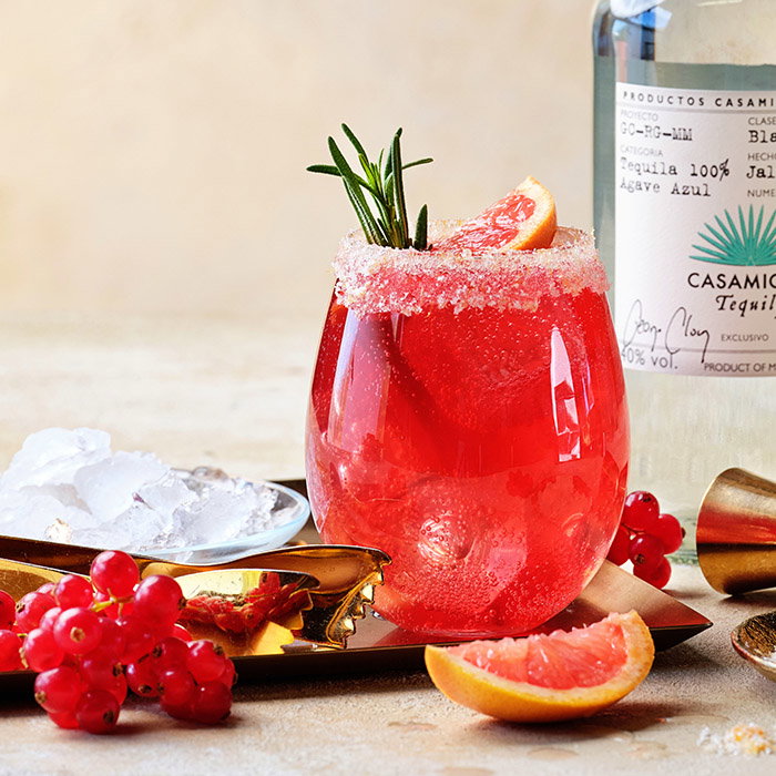 A vibrant cocktail with a sugar rim, garnished with grapefruit and rosemary, beside fresh berries and a bottle of tequila.