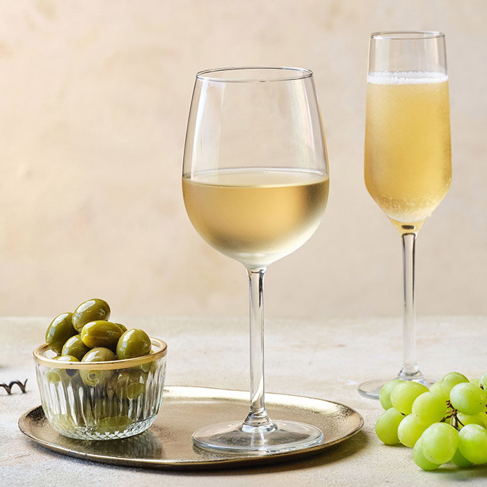 A glass of white wine and a flute of sparkling wine beside a bowl of green olives and fresh grapes on a gold tray.