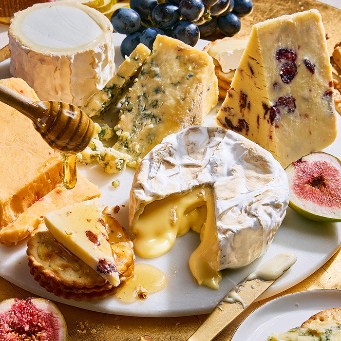 A variety of cheeses on a board, including a melted Camembert, blue cheese, and cheddar with cranberries. Honey drizzles over sliced crackers.