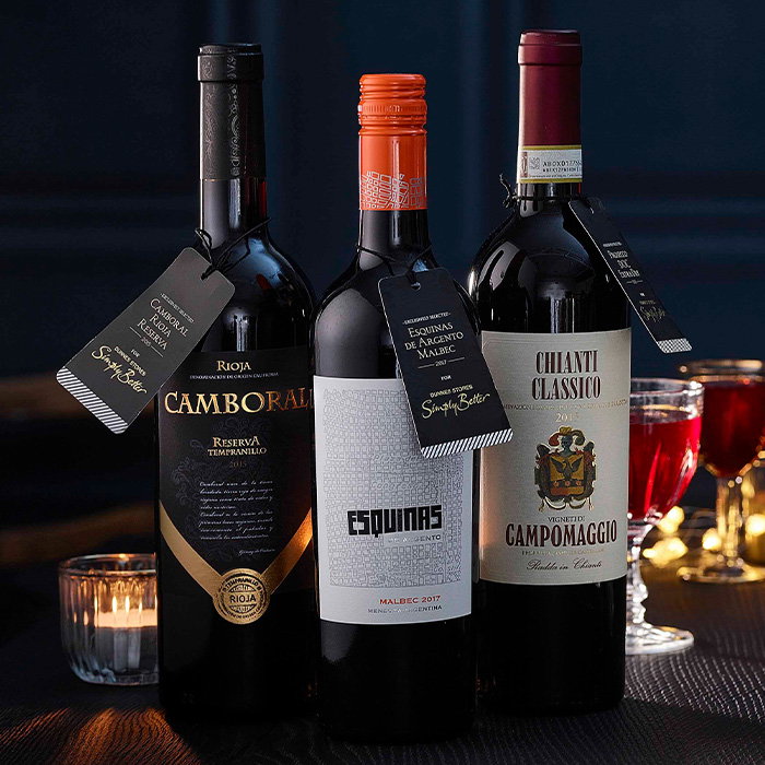 Three wine bottles with elegant tags, set against a dark background. Two glasses with red wine and a tealight candle add a festive touch.