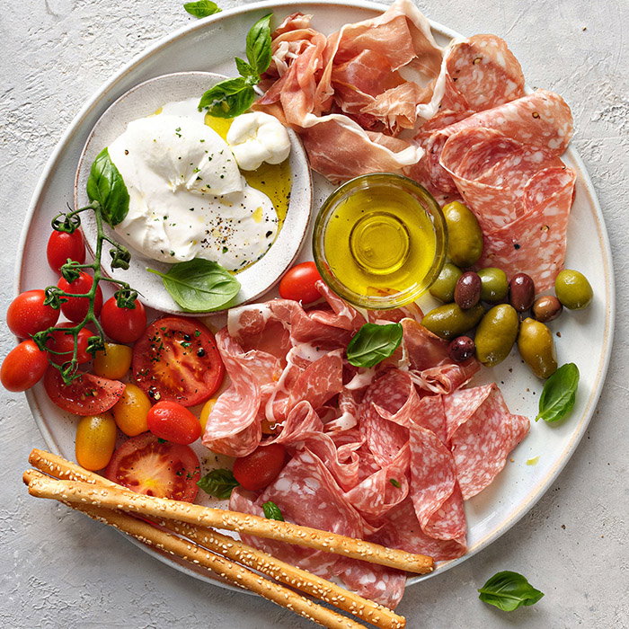 A platter featuring assorted cured meats, fresh tomatoes, olives, burrata cheese, basil, olive oil, and pretzel sticks.