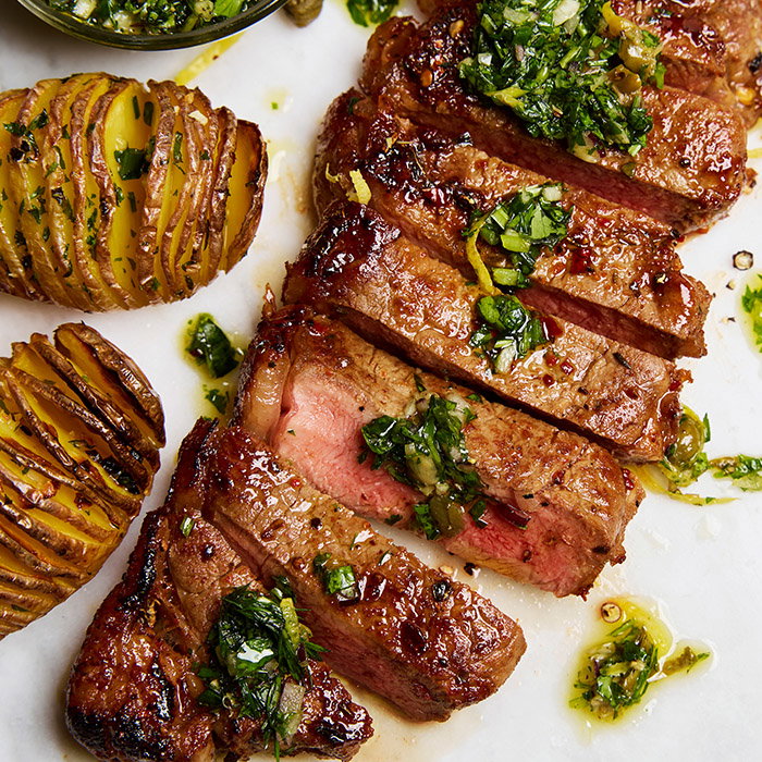 Grilled steak slices topped with herbs and sauce, alongside crispy, accordion-cut roasted potatoes on a white plate.