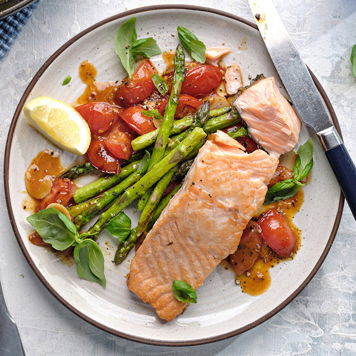 Grilled salmon fillet on a plate with asparagus, cherry tomatoes, fresh basil, and a lemon wedge. A knife rests on the plate. Inviting and fresh.