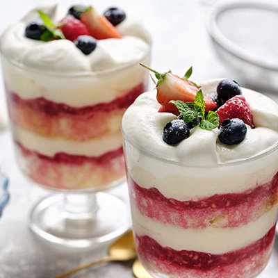 Two layered desserts in clear cups, featuring whipped cream, berry compote, and fresh strawberries and blueberries, garnished with mint.