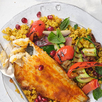 Plate of couscous topped with grilled fish, vibrant roasted vegetables, fresh herbs, and pomegranate seeds.