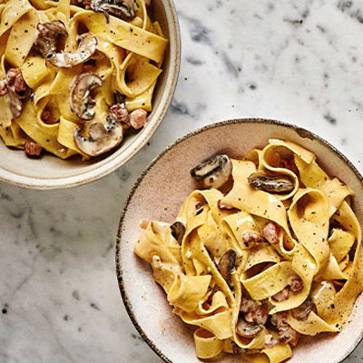 Two bowls of creamy mushroom and bacon fettuccine on a marble surface, garnished with black pepper.