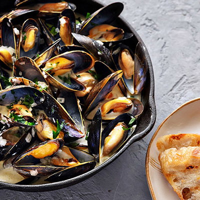 A cast-iron skillet filled with steamed mussels in a creamy sauce, garnished with parsley, alongside a slice of crusty bread.