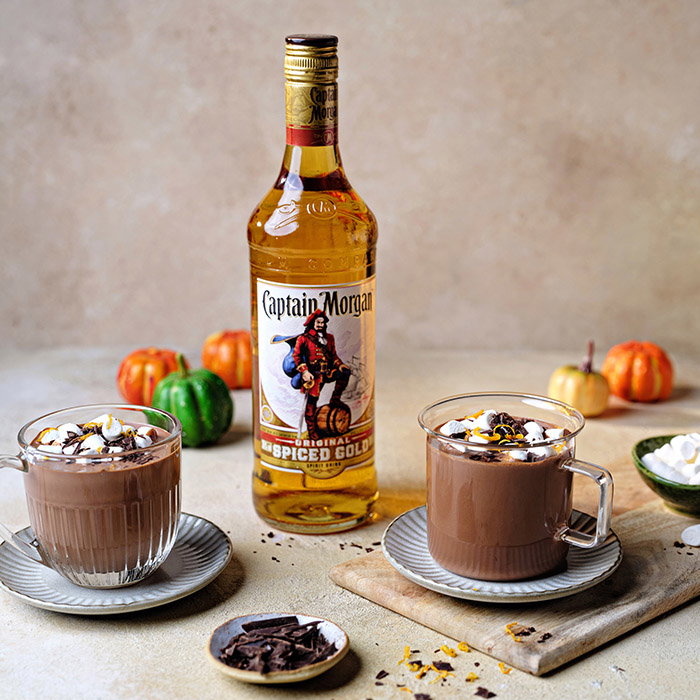 Two mugs of hot chocolate topped with marshmallows, beside a bottle of Captain Morgan, and surrounded by small pumpkins.