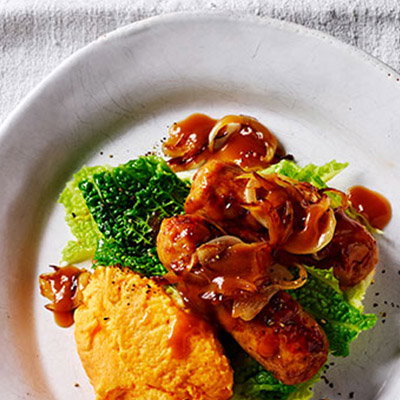 A plate featuring sausages topped with onion gravy, served with mashed pumpkin and wilted green cabbage.