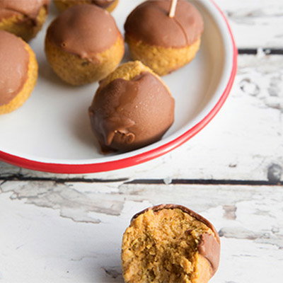 A plate of chocolate-dipped cake balls, one partially bitten showing a moist, golden interior. Speckled wooden tabletop background.