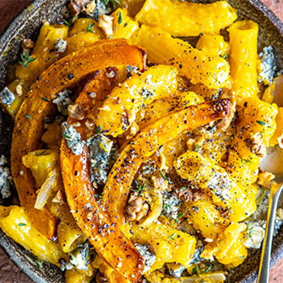 A close-up of creamy pasta with roasted squash, walnuts, and blue cheese, topped with fresh herbs and cracked black pepper.