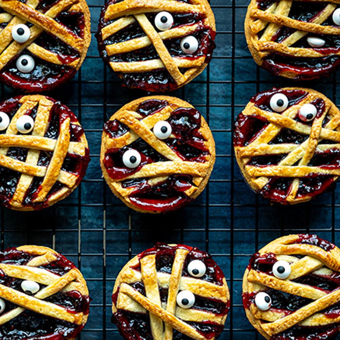 A top view of individual mummy-themed pies with lattice crust and candy eyes, cooling on a wire rack.