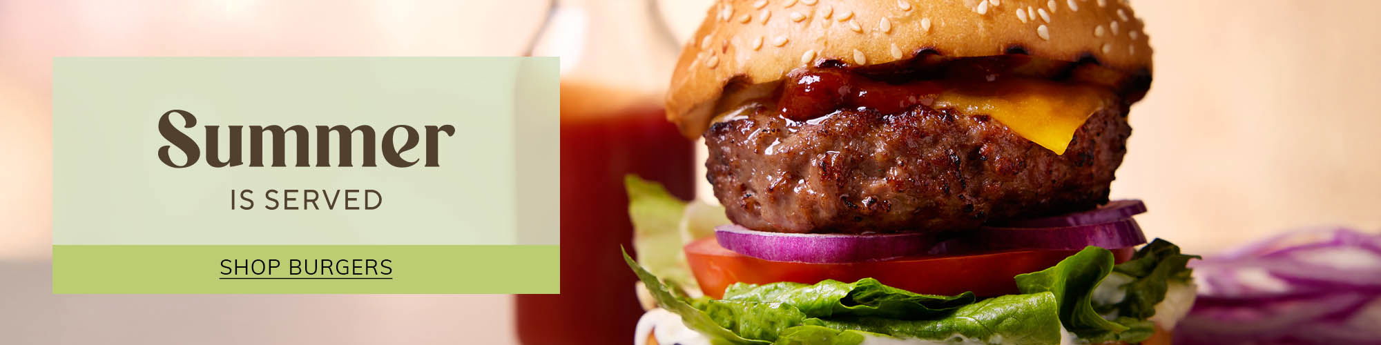 Shop all things burger. Cheeseburger with big beef patty, ketchup, lettuce, red onion, tomato, on a seeded bun and a ketchup bottle in the background out of focus.