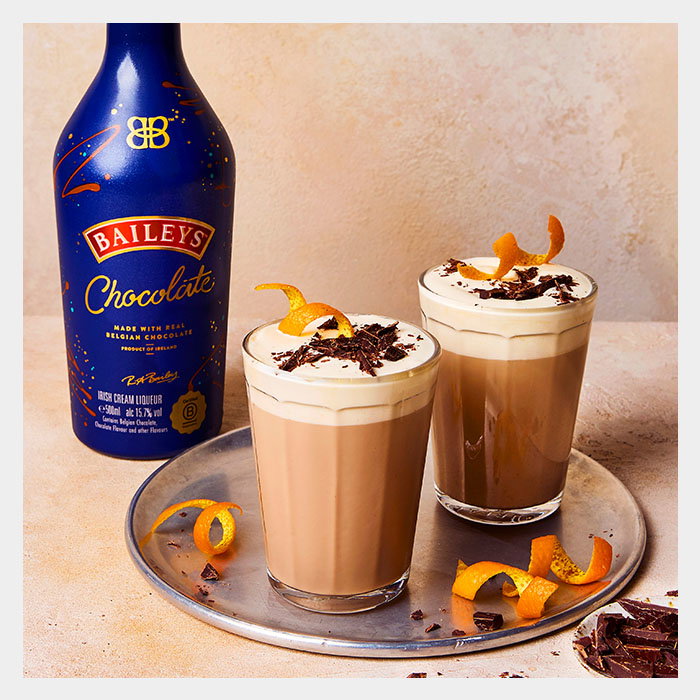 Two glasses of a creamy chocolate drink topped with whipped cream, chocolate shavings, and orange peels, beside a bottle of Baileys Chocolate.