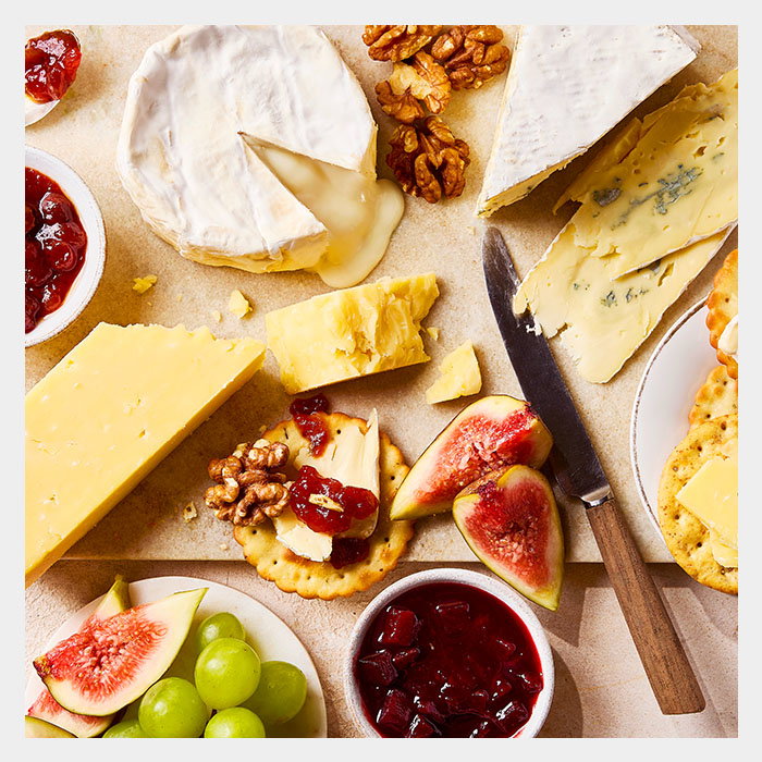 A cheese platter featuring various cheeses, nuts, figs, grapes, crackers, and bowls of jam, arranged on a wooden surface.