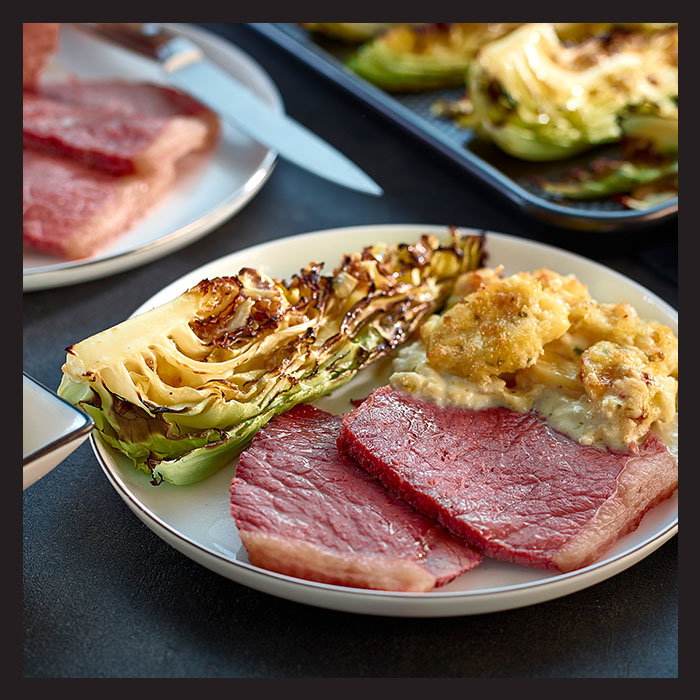 A plate with slices of corned beef, roasted cabbage, and cheesy cauliflower. The dish has a rustic, hearty appearance, suggesting warmth and comfort.