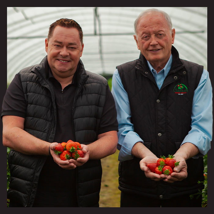 Neven Maguire and Pat Clarke, smiling, stand in a greenhouse holding bright red strawberries. They wear black vests, conveying a sense of pride and warmth.