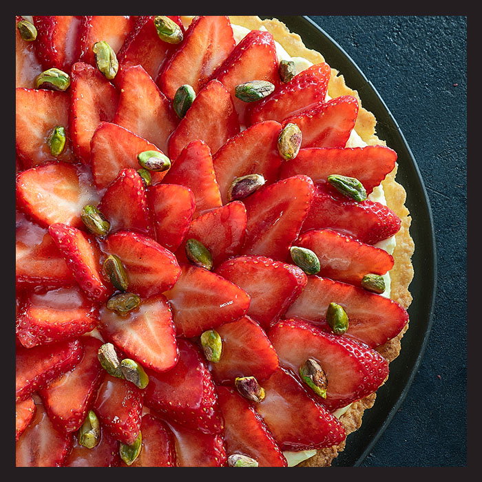 A vibrant strawberry tart is garnished with sliced strawberries arranged in circular layers, adorned with scattered green pistachios, creating a fresh, inviting look.