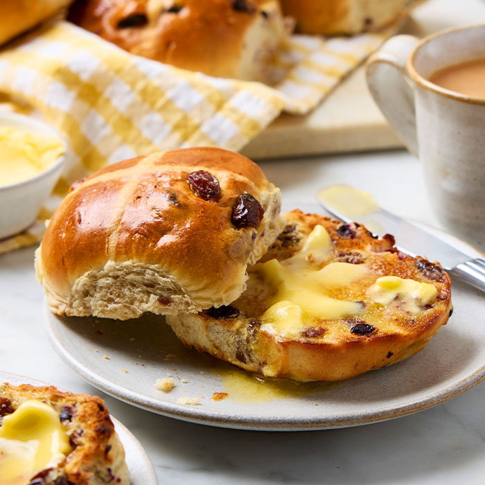 Toasted hot cross bun with melted butter on a plate, accompanied by a cup of tea and additional buns in the background.