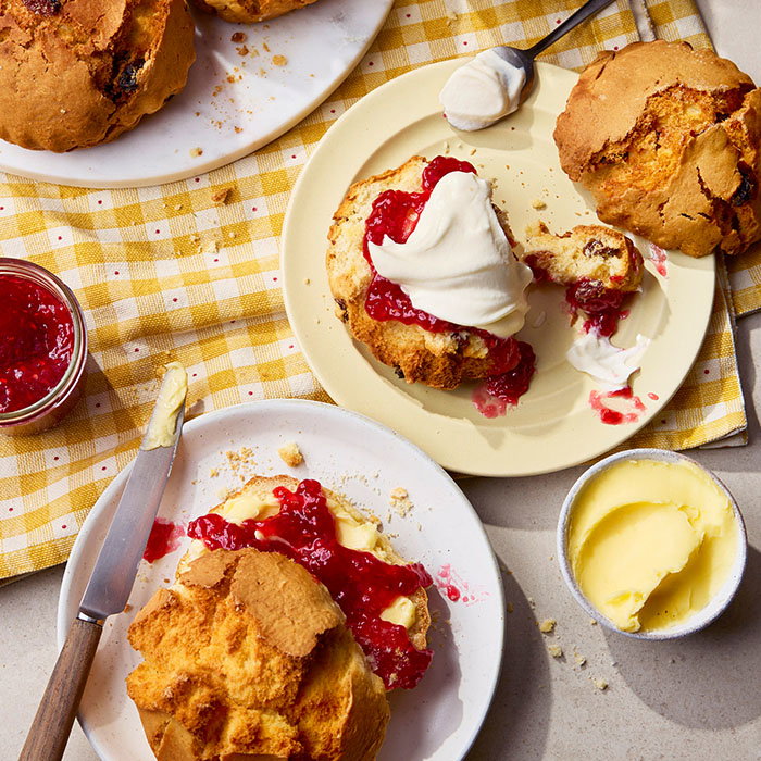Two plates with scones topped with clotted cream and red jam on a yellow checkered cloth, accompanied by a butter dish and a jar of jam.