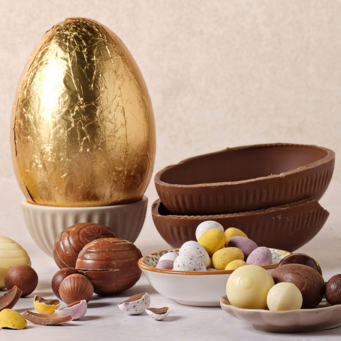 Assorted chocolate Easter eggs, including a large gold-wrapped egg, hollow milk chocolate halves, and colorful candy-coated mini eggs in bowls.