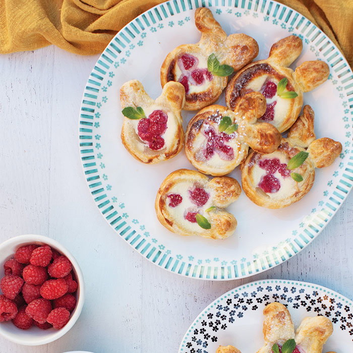 Plate of bunny-shaped raspberry pastries garnished with mint leaves, accompanied by a bowl of fresh raspberries on a white wooden table.