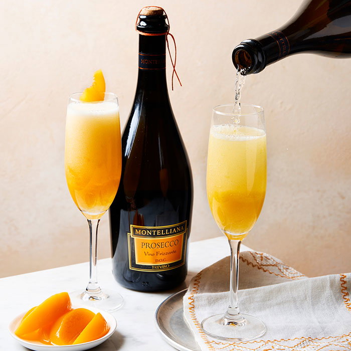 A bottle of Montelliana Prosecco with two peach Bellinis in champagne flutes, one being poured, next to a plate of peach slices and a folded napkin.