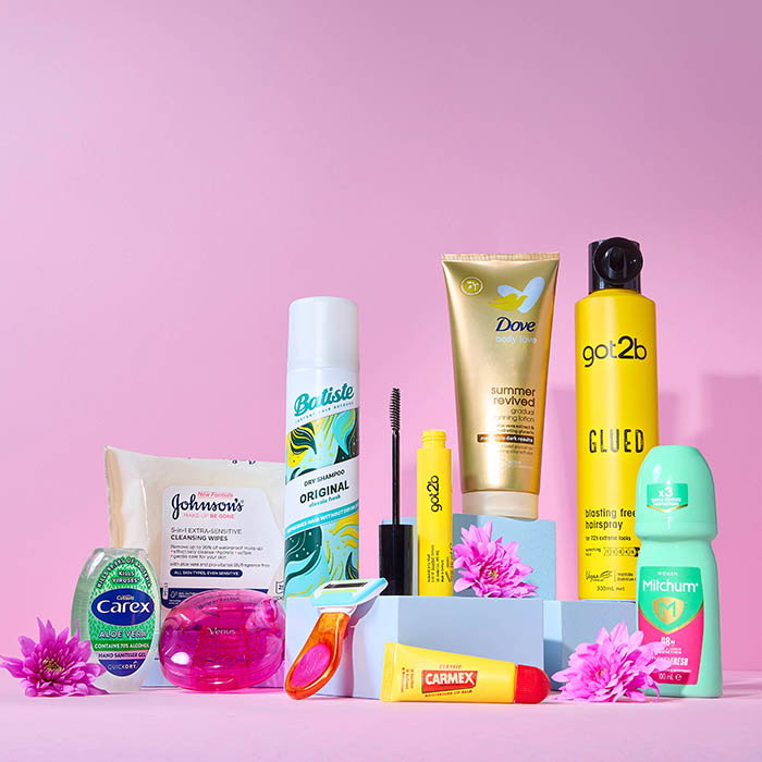 Variety of personal care products including dry shampoo, hair gel stick, deodorant, hand sanitiser, and wipes arranged with pink flowers on a pink background.
