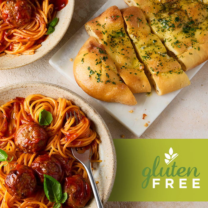 Plates of spaghetti with meatballs and basil, alongside sliced garlic bread on a white tray, labeled gluten free.