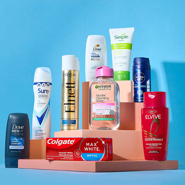 Various personal care products including deodorants, facial wash, cleansing water, toothpaste, and shampoo displayed on pink platforms against a blue background.