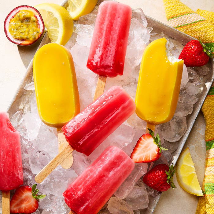 Tray of red and yellow fruit popsicles on ice, garnished with strawberries, lemon slices, and passion fruit half.