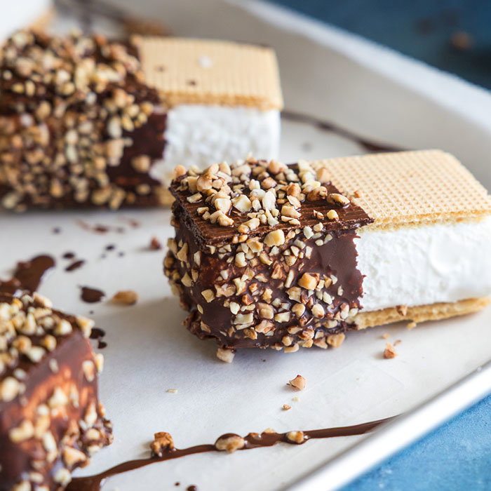 Wafer ice cream sandwiches dipped in chocolate and coated with chopped nuts on a white tray.