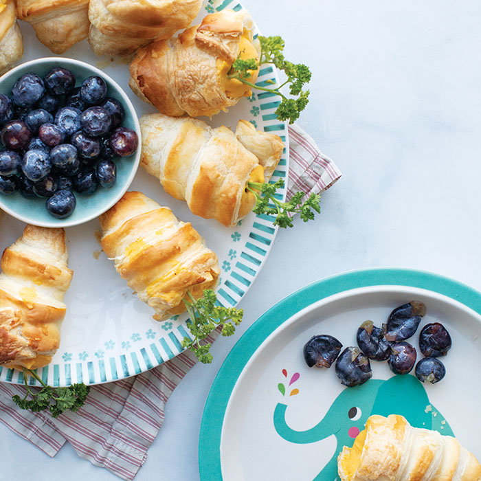 Plate of crescent rolls stuffed with cheese and parsley, a bowl of blueberries, and a children's plate with an elephant design holding crescent roll and blueberries.