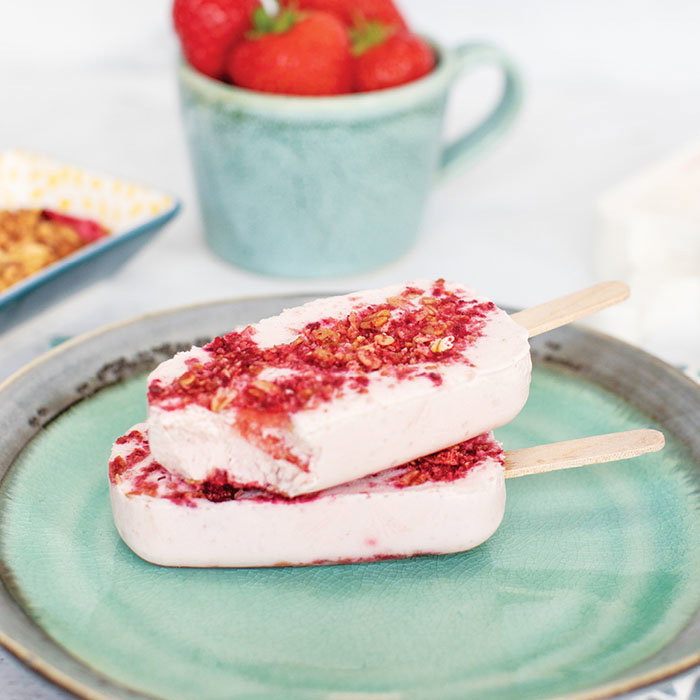 Two strawberry and granola popsicles stacked on a green ceramic plate with a mug of fresh strawberries in the background.