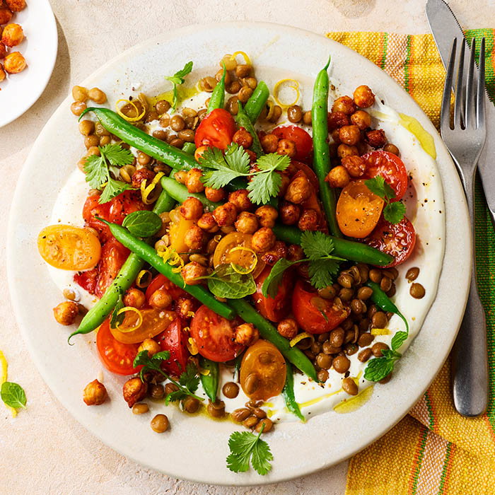 Plate of chickpea and lentil salad with cherry tomatoes, green beans, fresh herbs, and yogurt sauce, served with olive oil drizzle.