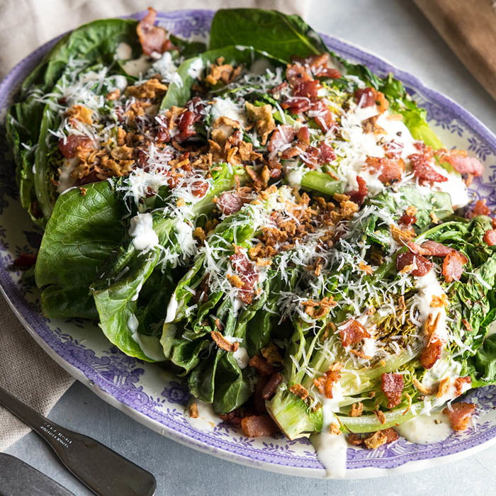 Grilled romaine lettuce salad topped with creamy dressing, crispy bacon bits, shredded cheese, and fried onions on a decorative plate.