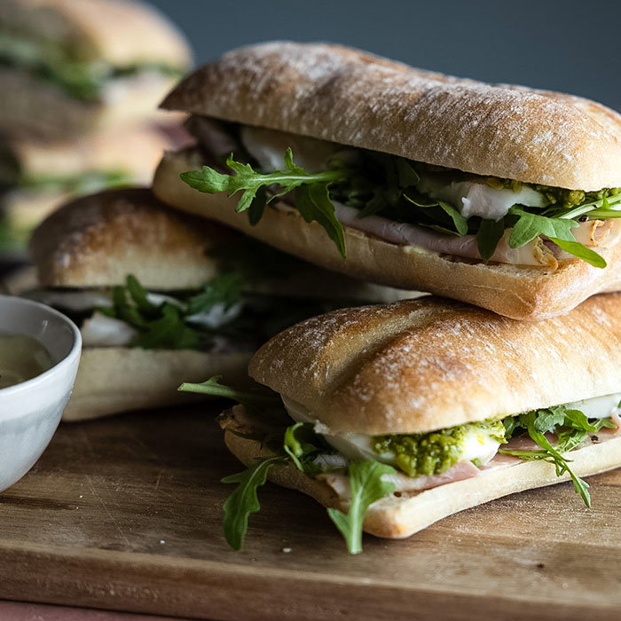 Four ciabatta sandwiches with ham, arugula, cheese, and green pesto on a wooden cutting board next to a small white bowl.