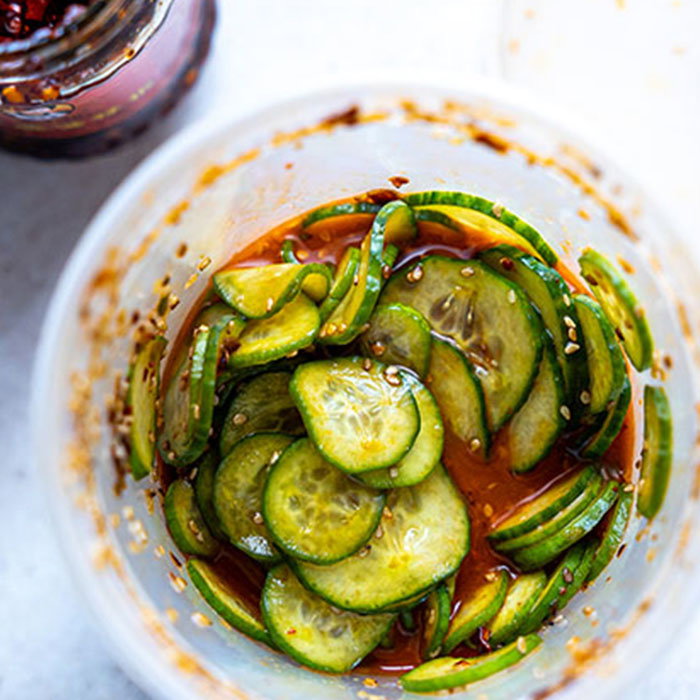 Pickled cucumber slices mixed with red chili oil and sesame seeds in a glass container on a white surface.