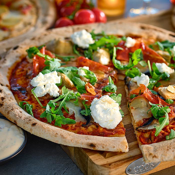 Thin-crust pizza topped with arugula, ricotta cheese, tomato sauce, mushrooms, and sliced cured meat on a wooden board.