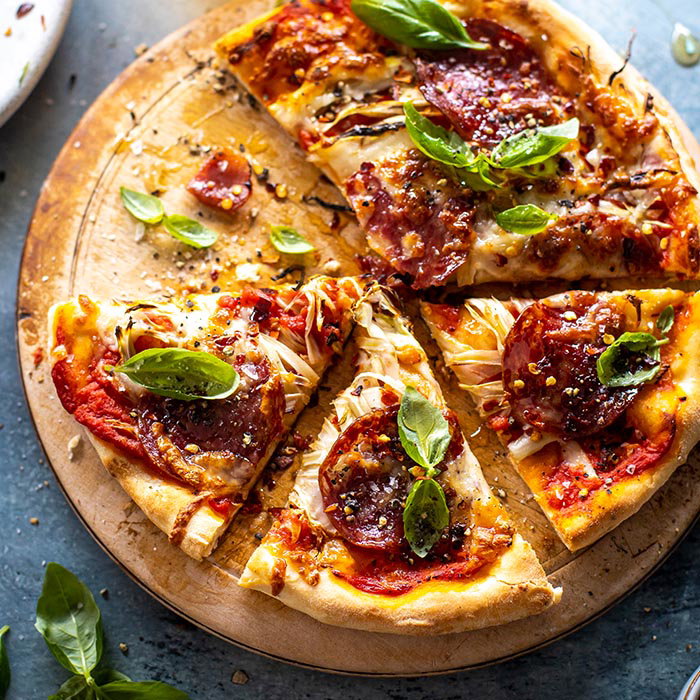 Sliced pepperoni pizza on a wooden board topped with fresh basil leaves, melted cheese, and red pepper flakes.