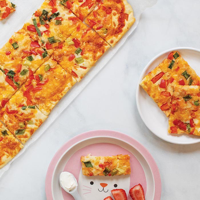 Rectangular cheesy vegetable pizza slices on white plates, with a cat-themed pink plate holding one slice and strawberries.