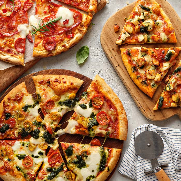 Three wooden boards with different pizzas: one with pepperoni, one with chicken and pesto, and one with cherry tomatoes and cheese.