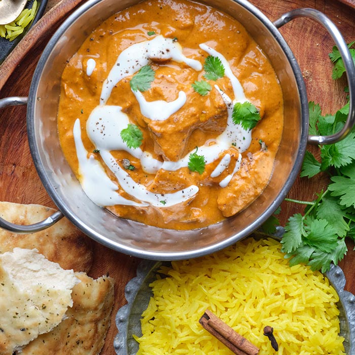 Top view of butter chicken garnished with cream and cilantro, served with yellow rice and naan bread on a wooden tray.