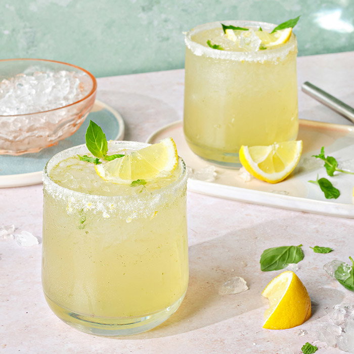 Two lemon cocktails with salted rims, garnished with lemon wedges and mint leaves, on a light pink surface with crushed ice.