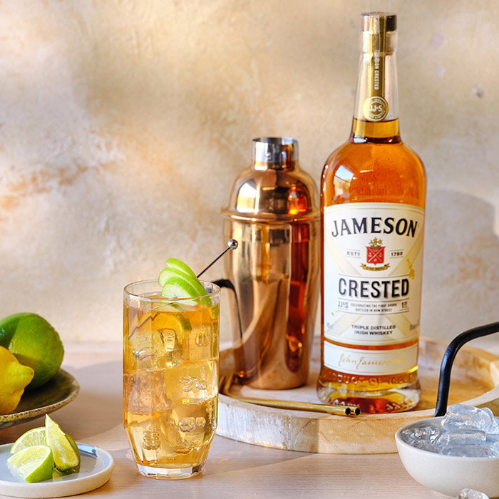 Bottle of Jameson Crested whiskey, cocktail with lime garnish, cocktail shaker, lime wedges, and bowl of ice on wooden surface.