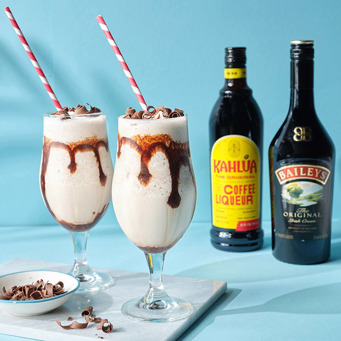Two creamy iced coffee drinks with chocolate garnish and striped straws, alongside bottles of Kahl&uacute;a coffee liqueur and Baileys Irish Cream.